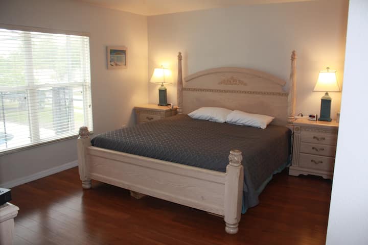 wide master bedroom with a large TV, dresser and 2 night stands, it also has a window overlooking the pool area.