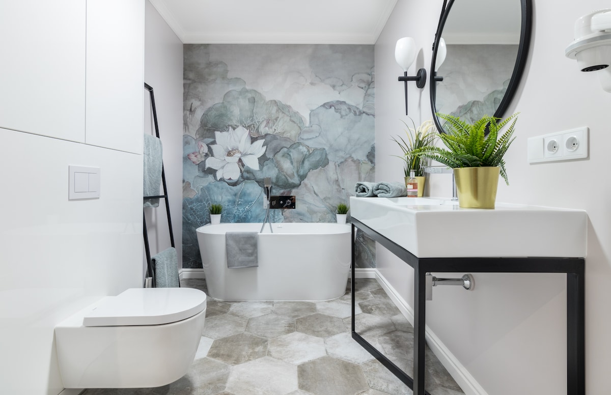 A modern bathroom is presented with a sleek freestanding bathtub and a well-lit vanity featuring a round mirror. A soothing floral mural adds color to the wall, while hexagonal tiles decorate the floor. Potted plants provide a touch of greenery throughout the space.