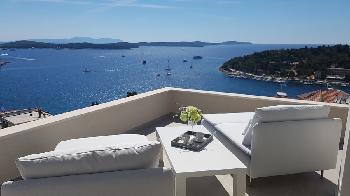A terrace is shown with two white lounge chairs and a small table, offering expansive views of the bright blue Adriatic Sea and distant islands. The sky is clear, and gentle waves are visible in the water, enhancing the serene ambiance of the space.