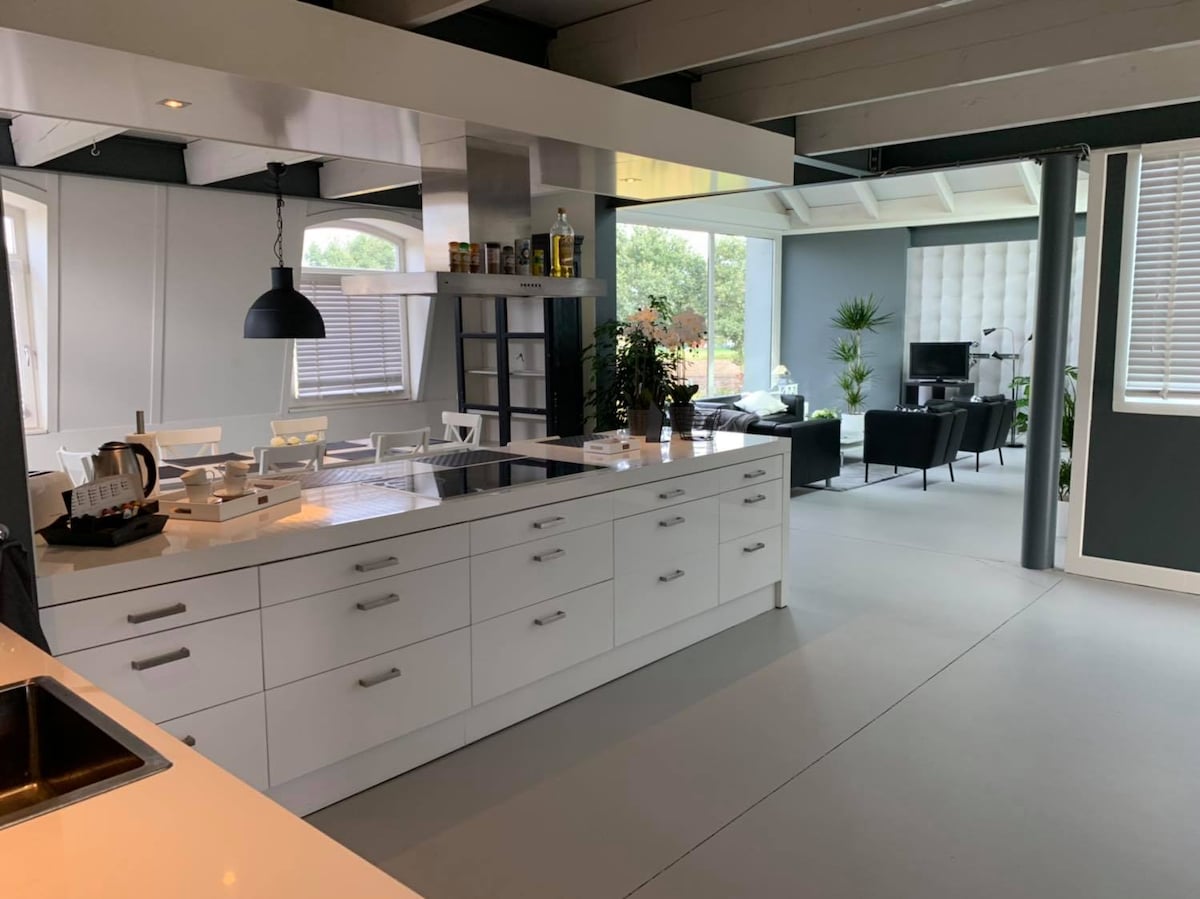 A modern kitchen features a central island with a stovetop and ample storage cabinets. Natural light streams in through large windows, illuminating the open layout that connects to a stylish living area. A sleek pendant light hangs overhead, adding sophistication to the space.