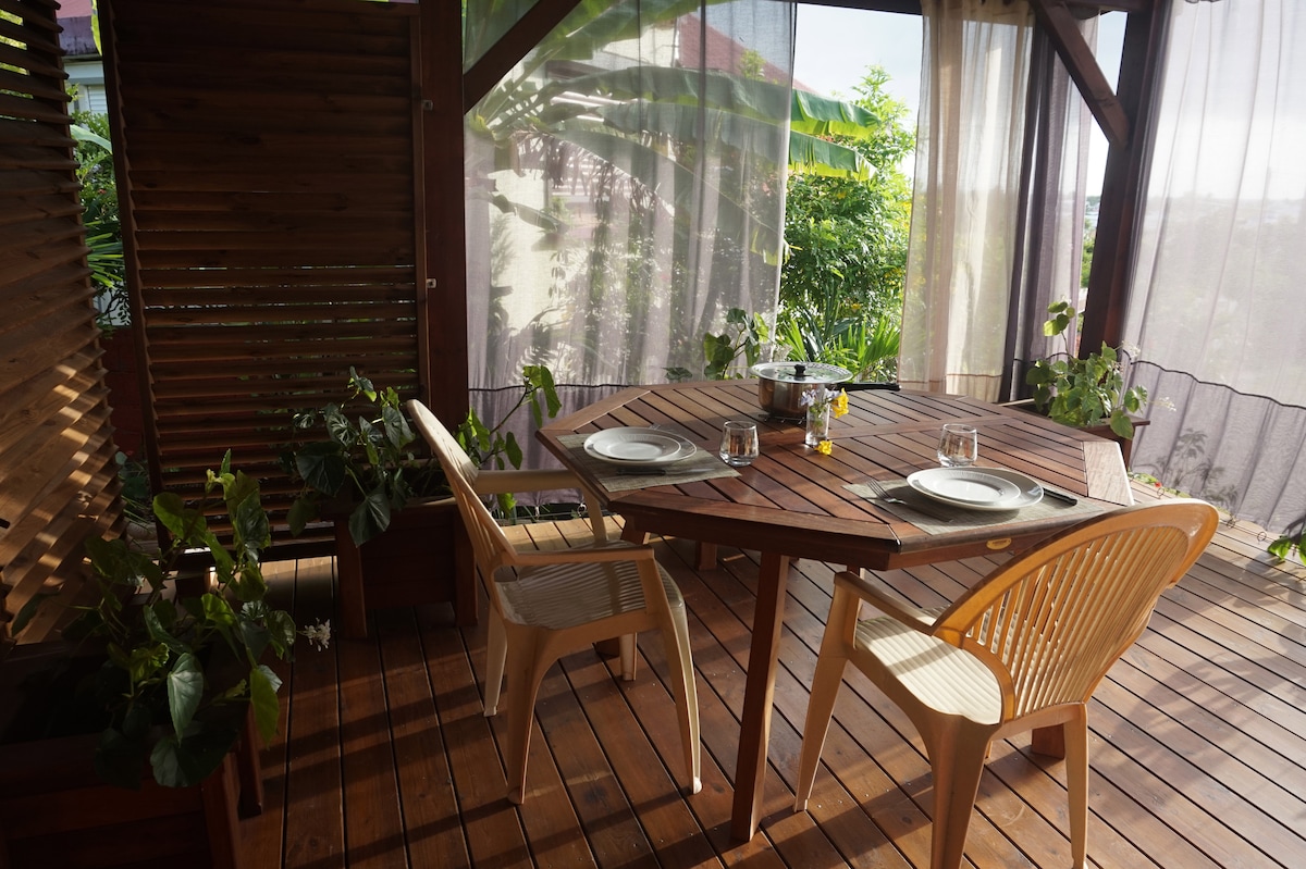 A private terrace features a wooden dining table surrounded by two contemporary chairs. Potted plants add greenery to the space, while sheer curtains provide a gentle filter to natural light. The setting is ideal for relaxed outdoor meals.
