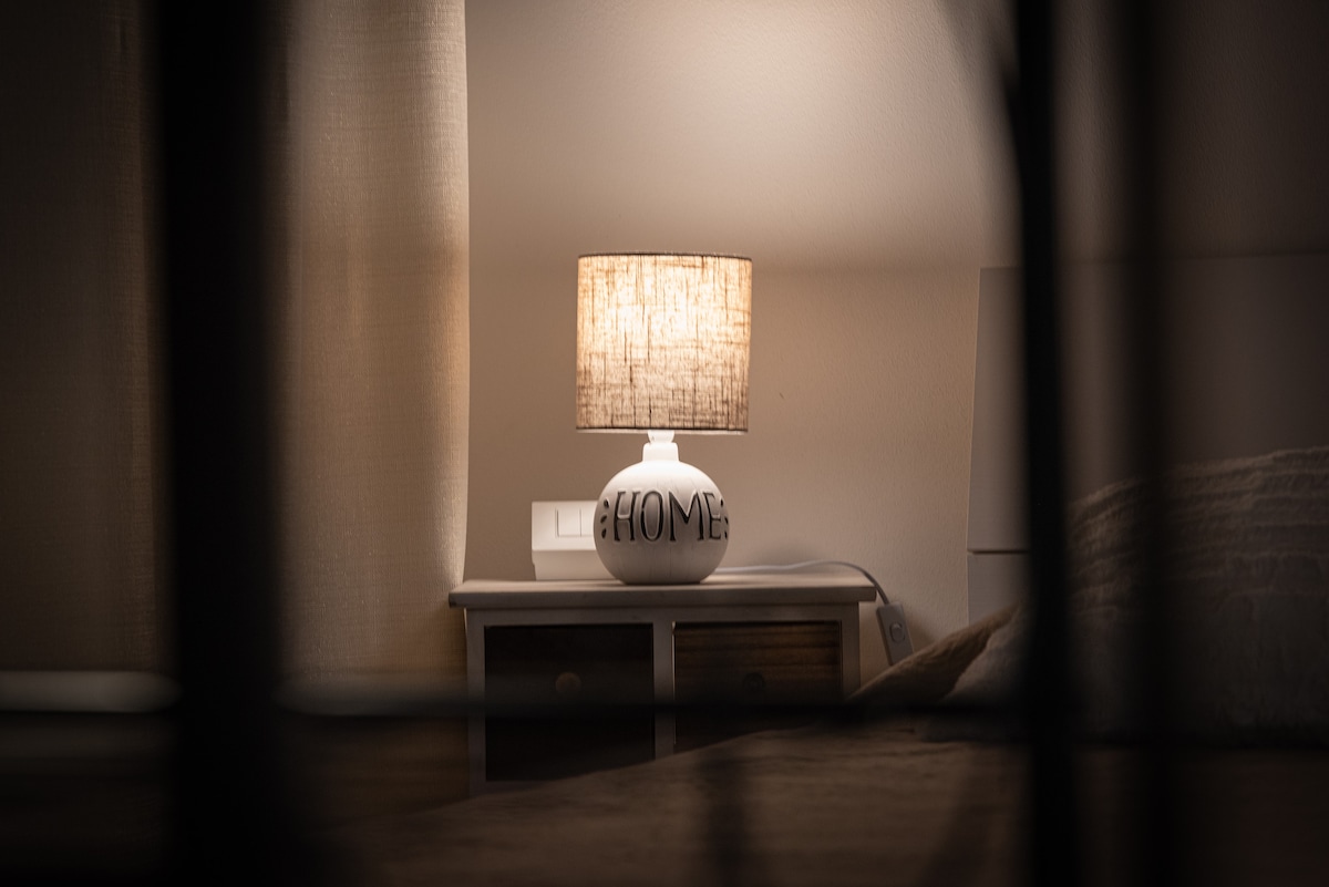 A bedside table is positioned next to a spacious bed, featuring a lamp with a fabric shade that casts a soft glow. The muted tones of the room are complemented by light curtains, creating a serene and restful environment.