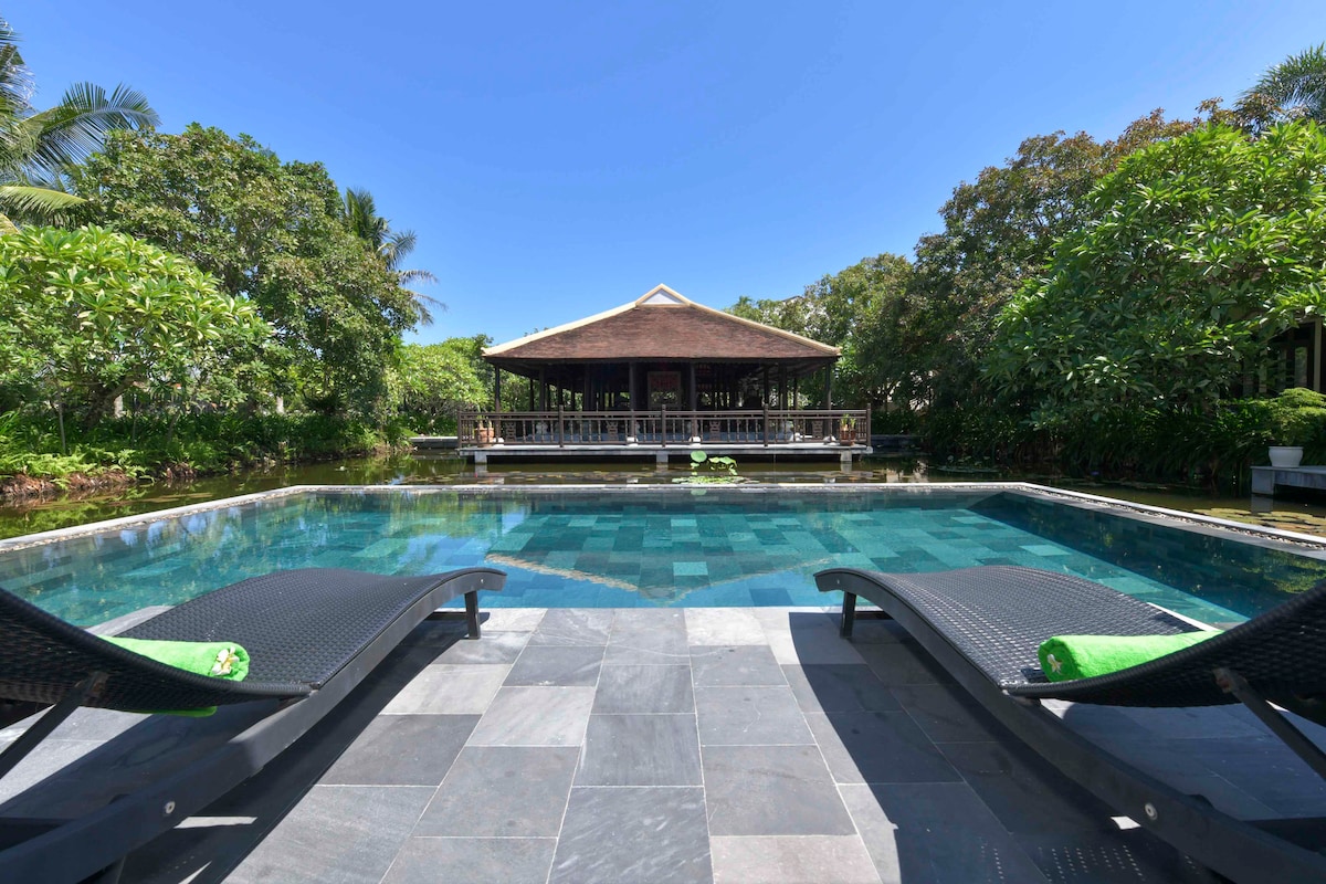The wet edge swimming pool is framed by lush greenery and features two sun loungers equipped with towels. A traditional wooden pagoda is visible in the background, set against a clear blue sky, creating a serene outdoor atmosphere.