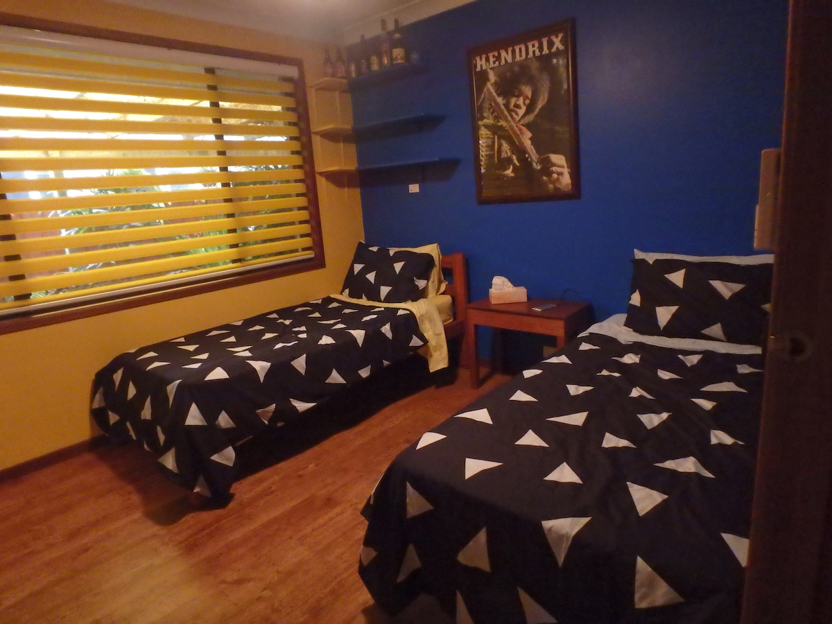 The twin share room features two beds with patterned bedding, positioned against a vibrant blue wall. A wooden bedside table and shelf are present, while light filters through the slatted windows, offering a view of the surrounding greenery.