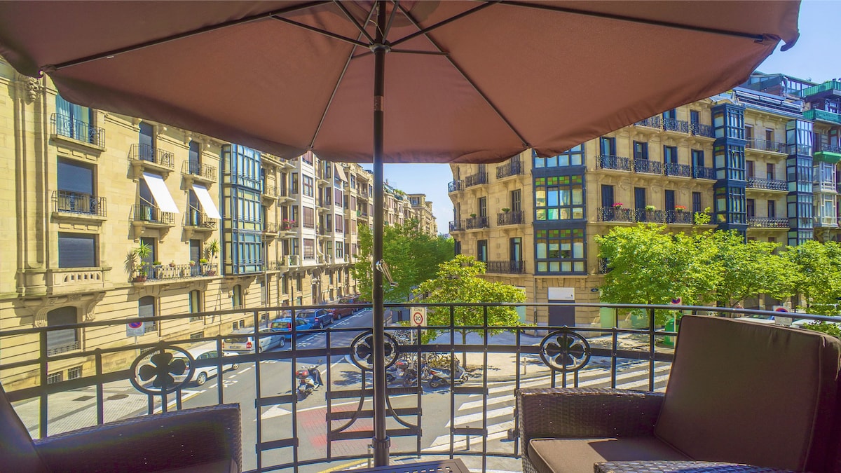 A balcony view captures the surrounding cityscape, featuring charming buildings with balconies and green trees lining the streets below. Two chairs are positioned on the balcony beneath a large umbrella, providing a comfortable spot to enjoy the scenery.