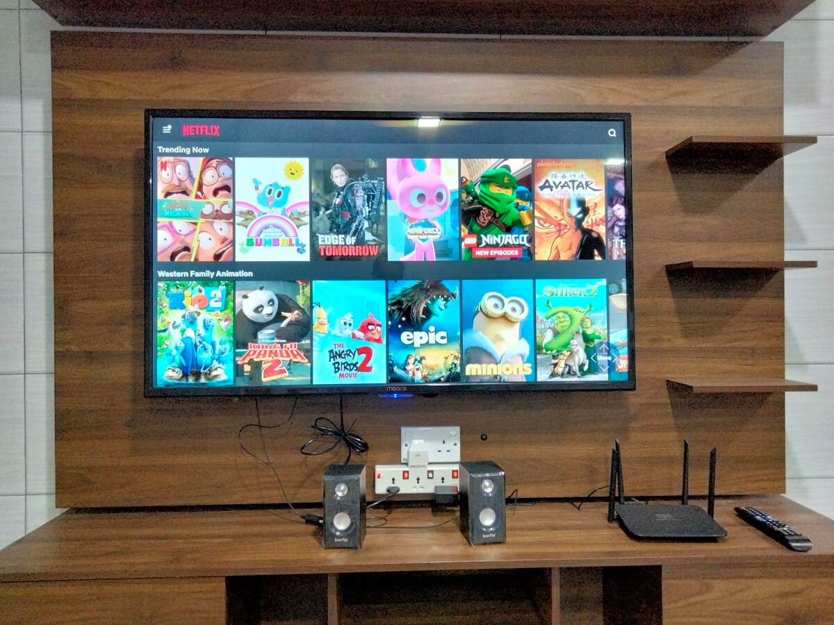 A large flat-screen television is mounted on a wooden wall unit, displaying various animated shows and movies available on a streaming platform. Below, a console holds speakers and a router, creating a convenient media setup.