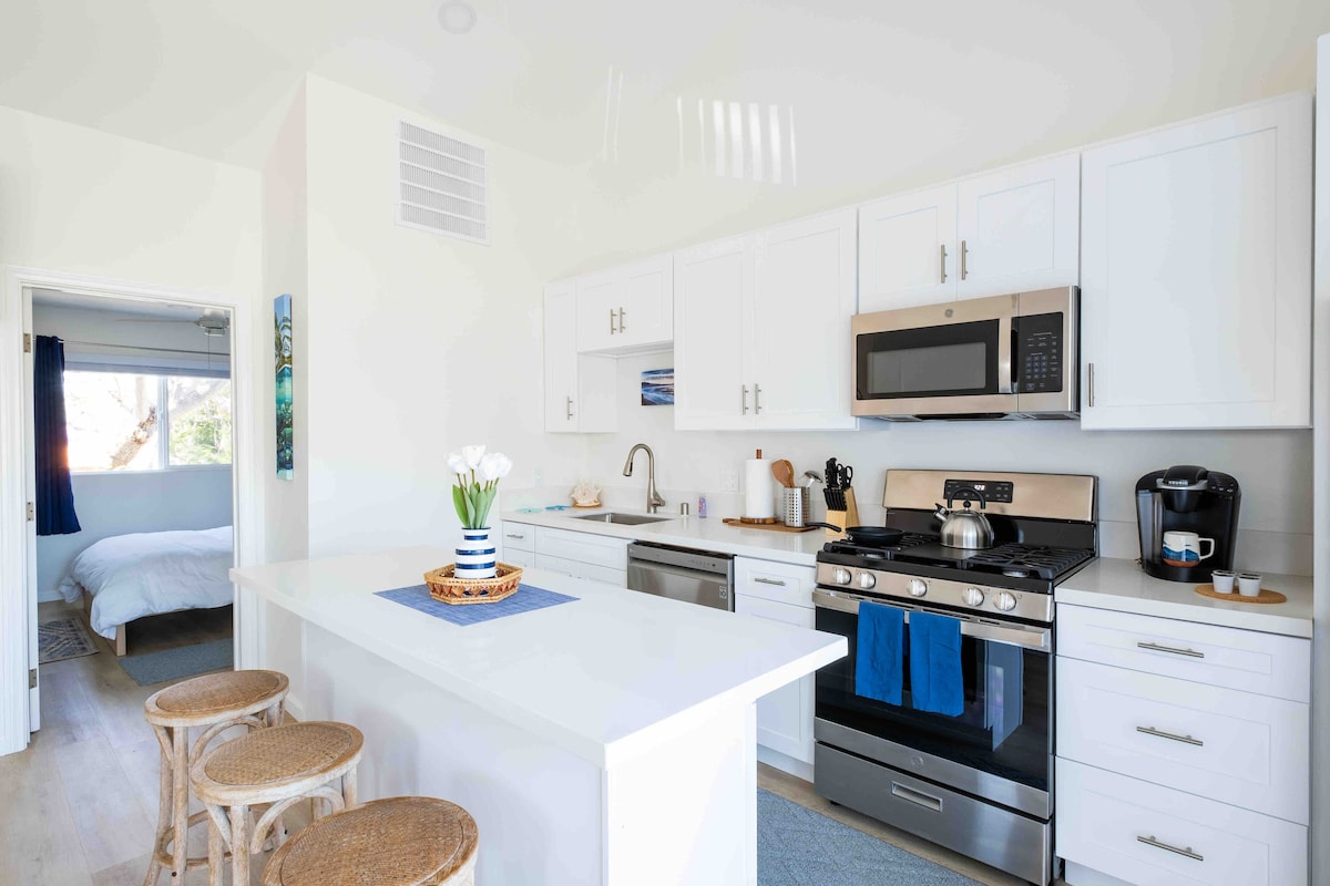 The kitchen features modern white cabinetry and stainless steel appliances. A large island provides additional counter space, with seating offered by three woven barstools. A colorful artwork is displayed on the wall, and a view into the adjoining sleeping area is seen through an open doorway.