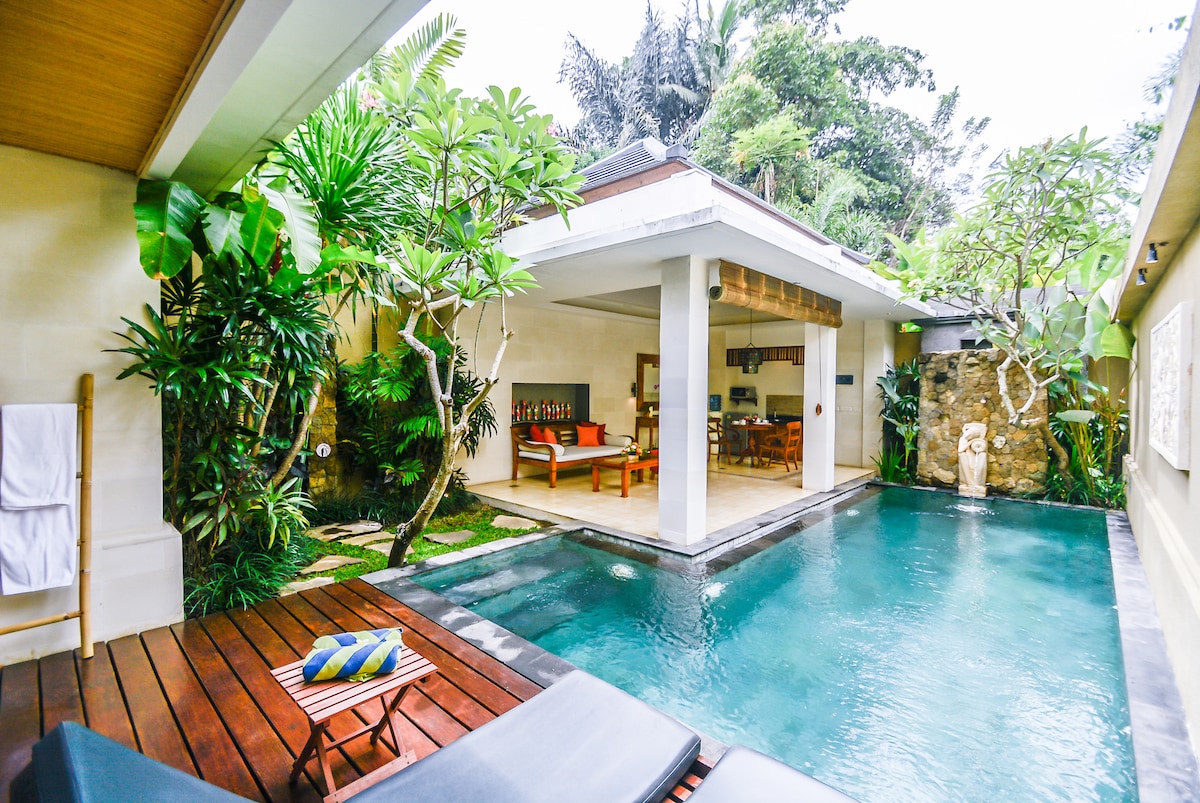 A private villa yard is viewable, featuring a sparkling pool surrounded by tropical greenery. Comfortable lounge chairs are positioned on a wooden deck, inviting relaxation. An open living area is seen through glass doors, with plush seating and dining space visible beyond the pool.