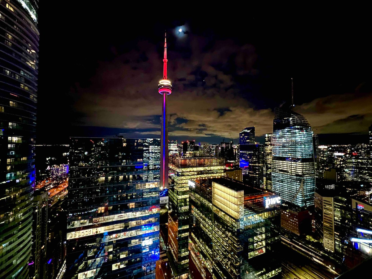 The city skyline is illuminated at night, featuring the CN Tower prominently lit in red. A mix of modern skyscrapers reflects various colors, with a clear sky and moon visible in the background, highlighting the vibrant atmosphere of Toronto.