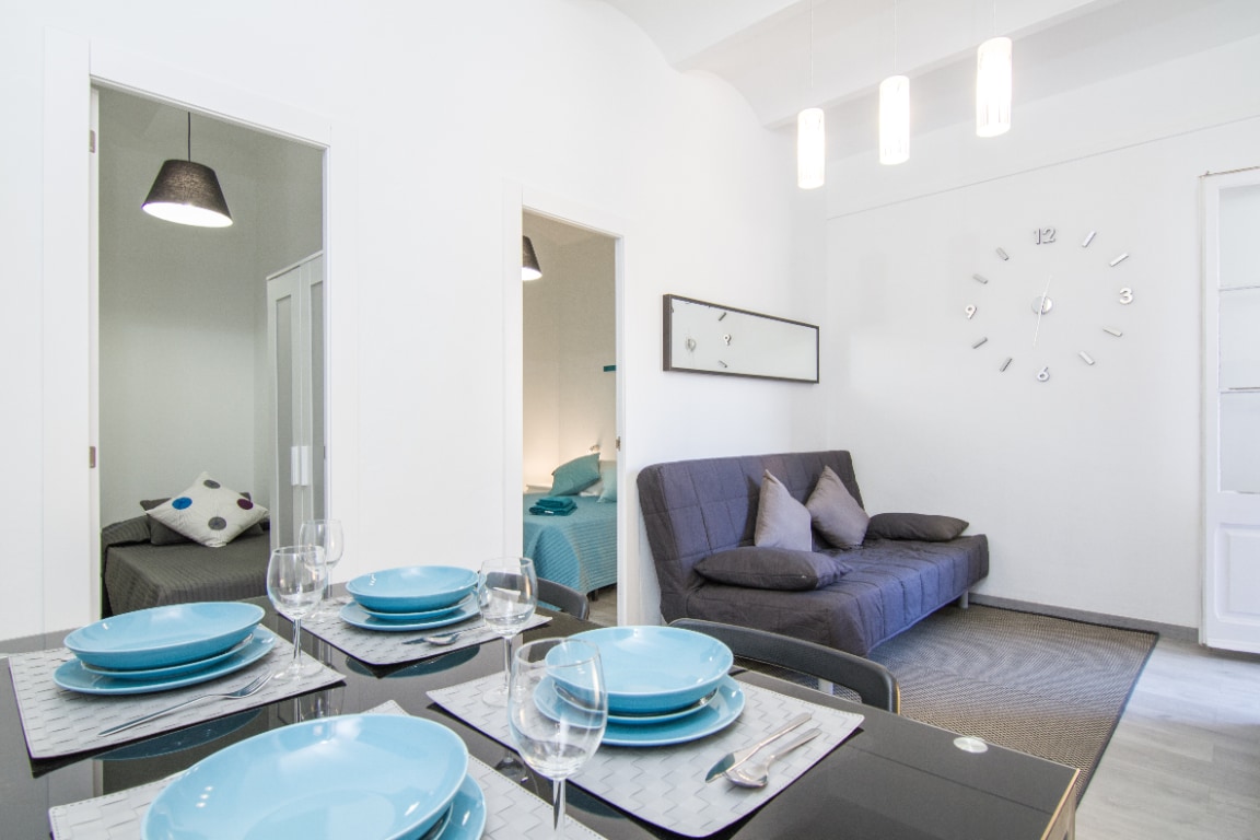 A bright and open living area includes a dining table set with blue plates and glasses. A sofa bed with contrasting cushions is positioned nearby, and a wall clock adds visual interest. Two doorways lead to separate sleeping areas, enhancing the apartment's functionality.