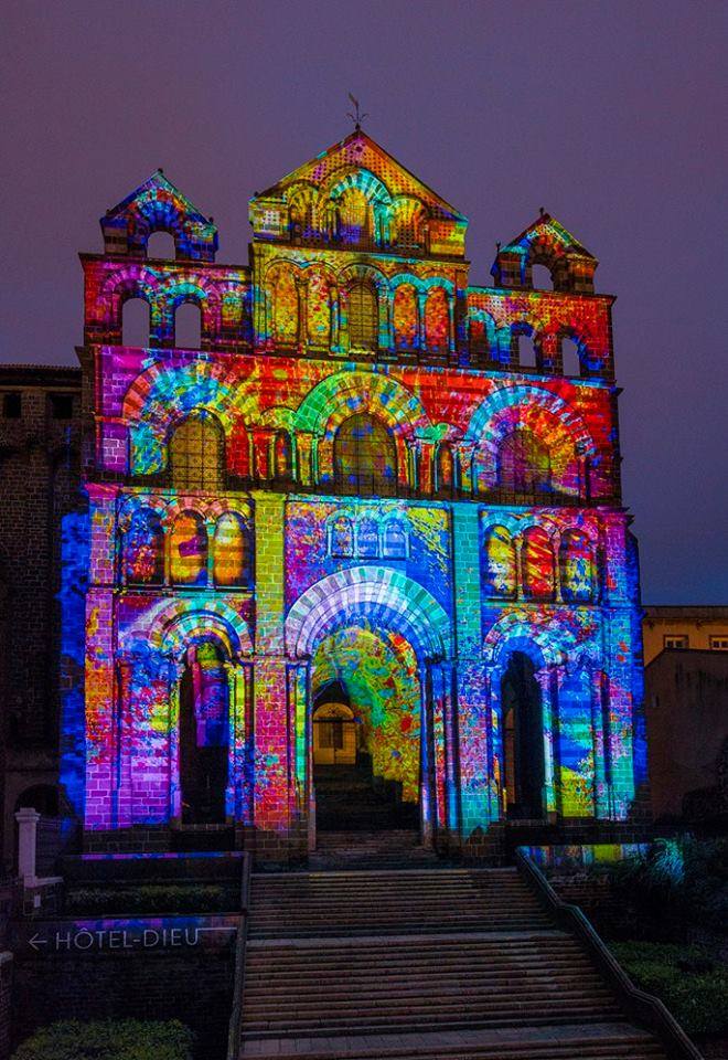 A historical building serves as a canvas for colorful projections, showcasing intricate patterns and vibrant hues. The illuminated facade highlights architectural details, while a staircase leads up to the structure. The scene captures a blend of heritage and artistry against a twilight backdrop.