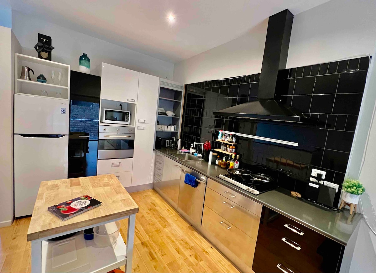 A modern kitchen features sleek stainless steel appliances, including an integrated oven and a large cooktop. Ample storage is provided by white cabinetry. A wooden island stands at the center, with a colorful decorative plate on top. Natural light illuminates the space.