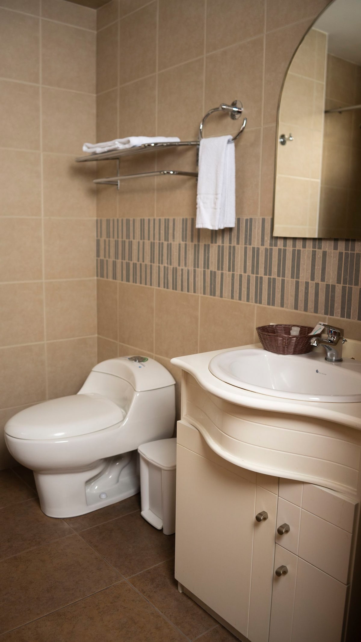 A modern bathroom is featured with neutral-toned tiles and a large mirror above the sink. A compact toilet is positioned beside a small waste bin. Shelving is installed above the sink, displaying clean towels.