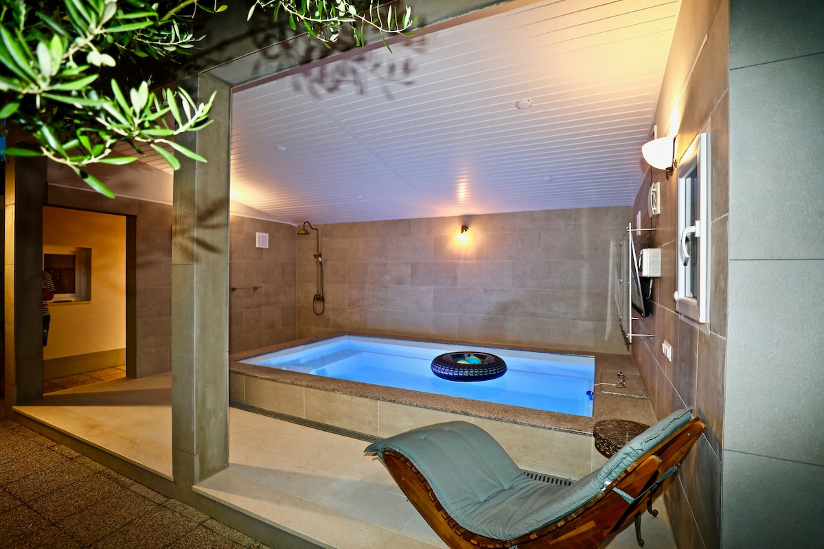 A private heated pool is set within a stone-walled space, illuminated by soft lighting. A comfortable lounge chair is positioned nearby, adding to the serene atmosphere. The pool water appears tranquil and inviting, complemented by a subtle view of olive tree leaves in the corner.