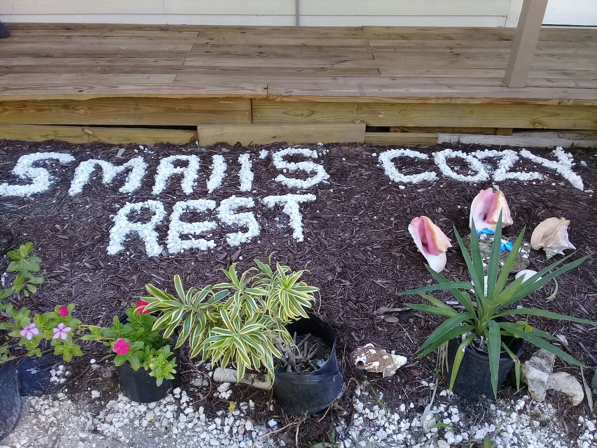 A welcoming outdoor space features a mulch area with the words 'Small's Rest' arranged in white stones. Several conch shells are placed nearby, alongside potted plants, including a pink flowering plant and greenery, contributing to the inviting appearance of the entryway.