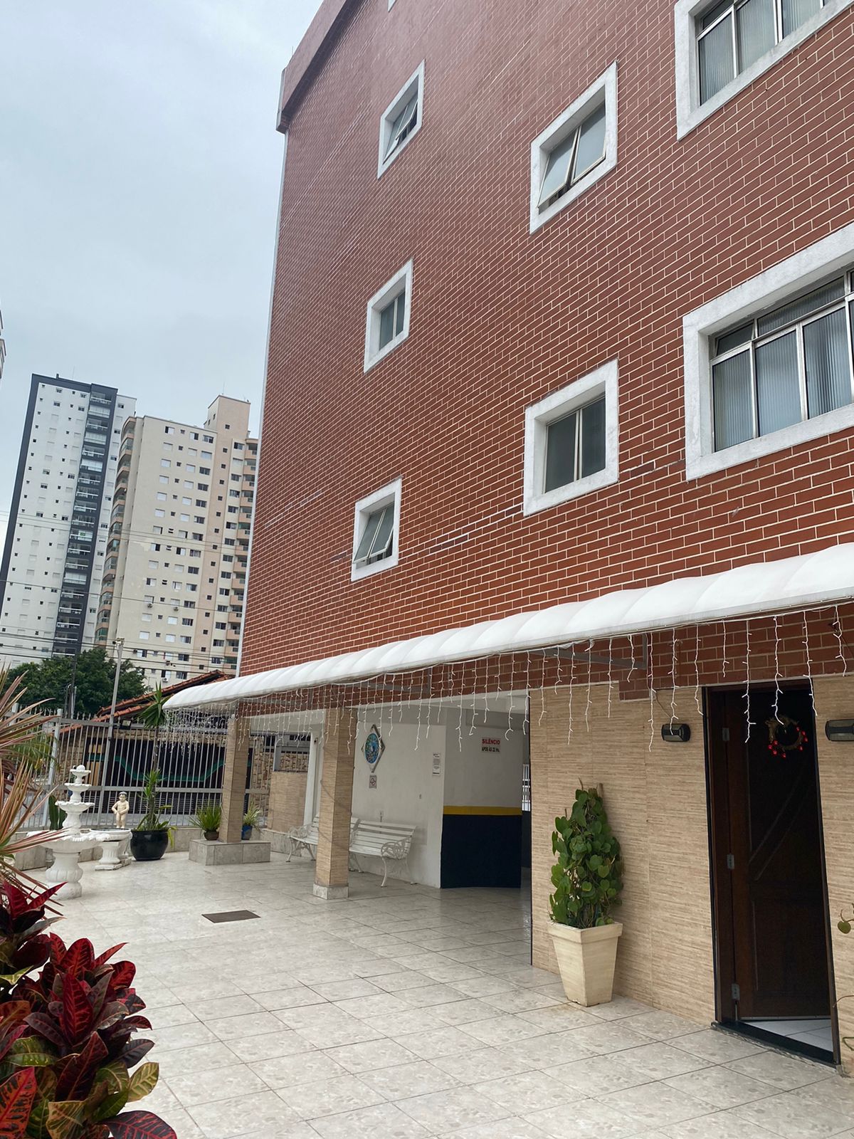 The exterior of a brick building is shown, featuring large windows and a covered entrance. The area is paved with tiles and includes decorative plants. Surrounding high-rise buildings are visible in the background, contributing to a lively urban setting.