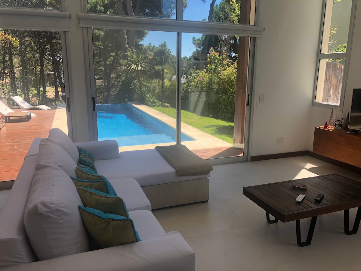 A bright living area features a white sectional sofa adorned with colorful cushions, positioned near large glass doors that open to an outdoor space. A wooden coffee table sits in front, with a serene view of a pool and garden visible through the glass.