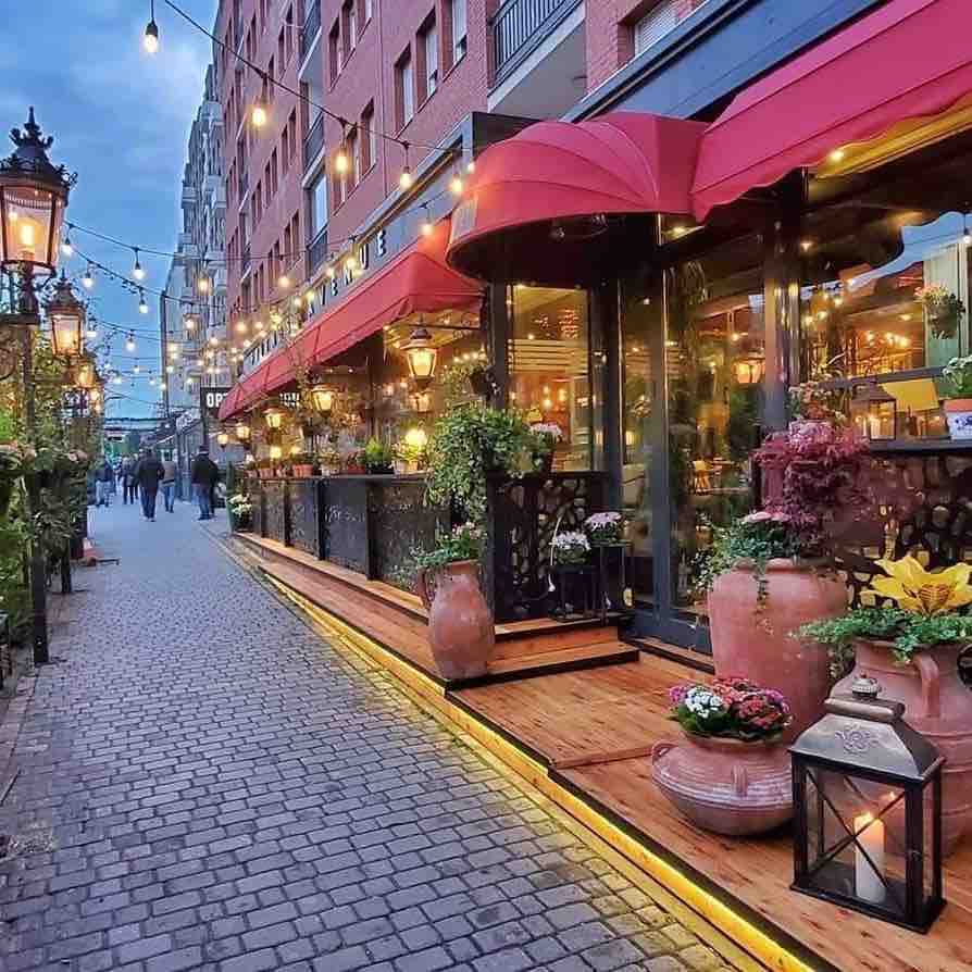 The street is lined with vibrant outdoor seating beneath red awnings, accompanied by warm string lights. Potted plants and decorative lanterns enhance the welcoming ambiance, while cobblestone paving leads past the café entrance, inviting passersby to explore the lively atmosphere of the area.