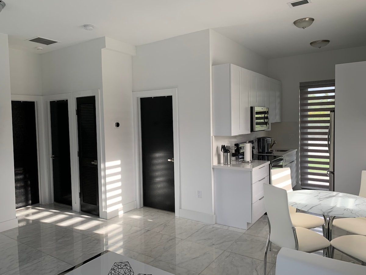 An open-concept living area is presented with marble flooring and modern cabinetry. Two sleek, black doors lead to separate rooms. A dining table with white chairs is positioned beside a large window, allowing natural light to fill the space through partially closed shutters.