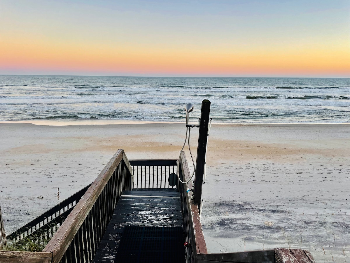 A wooden staircase leads down from the covered deck to the sandy beach, framed by a spacious outdoor shower on the left. The horizon showcases a gentle gradient of pastel colors at sunset, reflecting over the calm Atlantic Ocean.