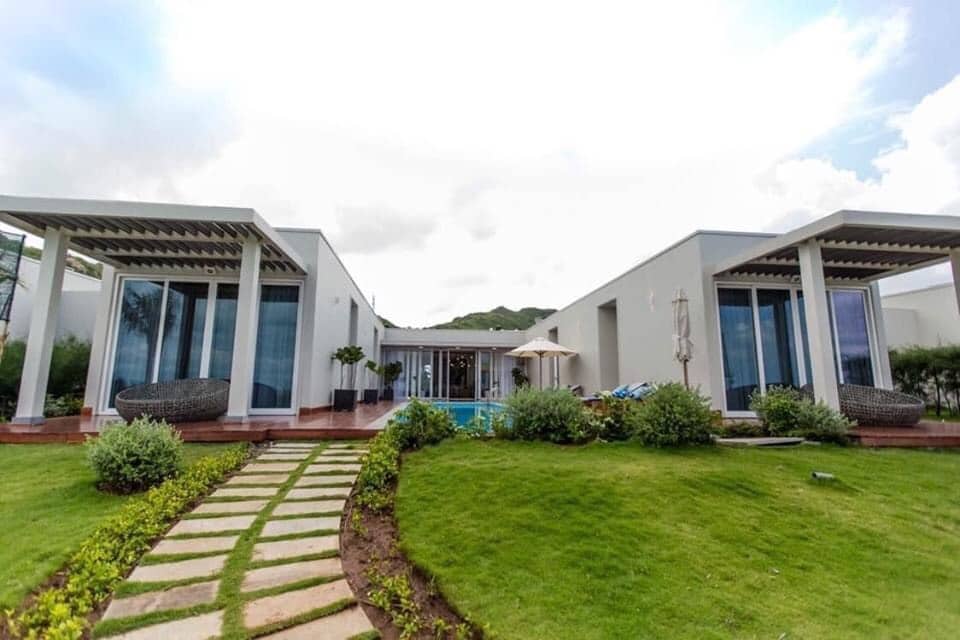 The villa is flanked by lush green lawns and neatly trimmed shrubs. A path lined with stepping stones leads to the entrance. The structure features large glass windows, offering views of the outdoor area, which includes a tranquil pool and shaded seating.