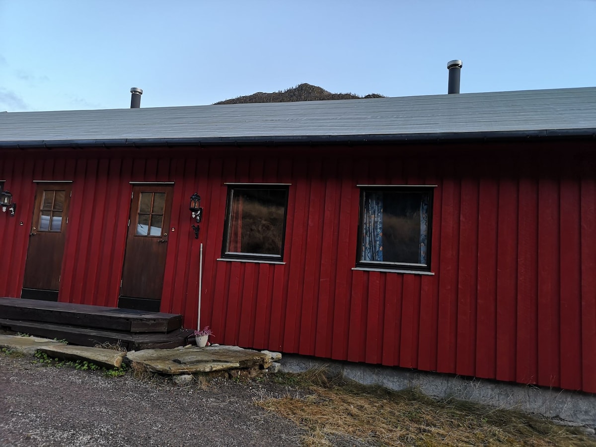 The exterior of a red wooden building is visible, featuring two doors and two windows. A wooden step leads to the entrance. The surrounding area shows a natural landscape with rocky terrain and vegetation. Soft light reflects off the building's surface.