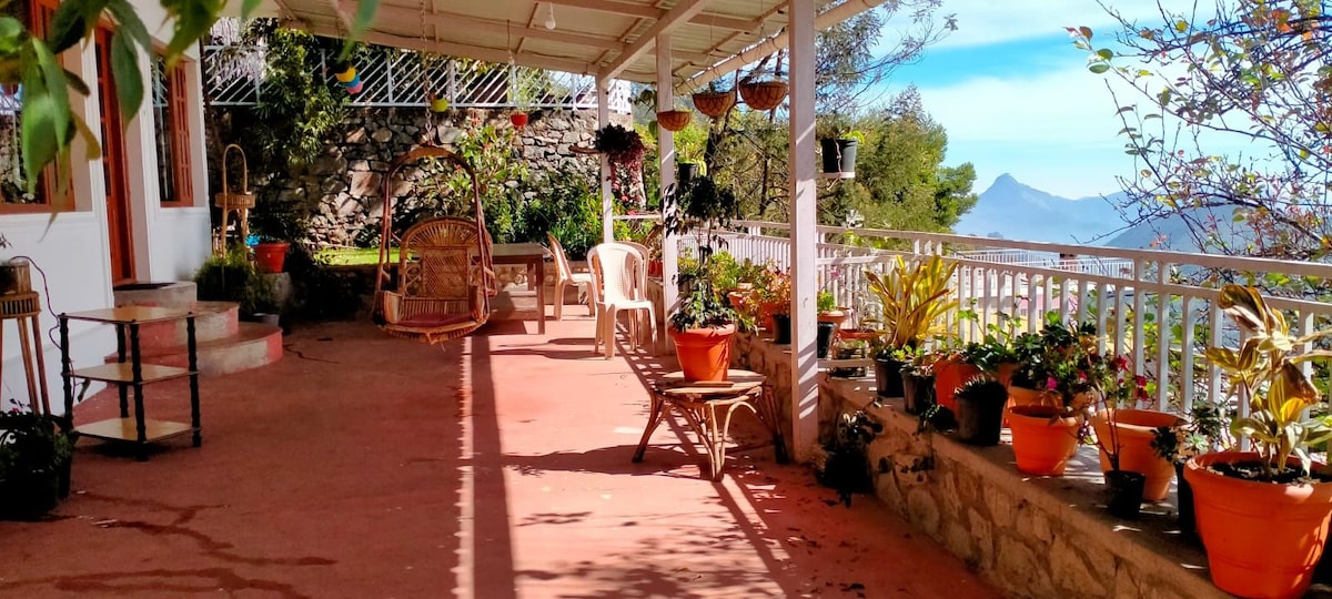 A spacious terrace is adorned with potted plants and hanging flower baskets. Seating options include a wooden swing and a bench. The area is shaded by a roof, allowing for relaxation while enjoying views of the surrounding landscape.