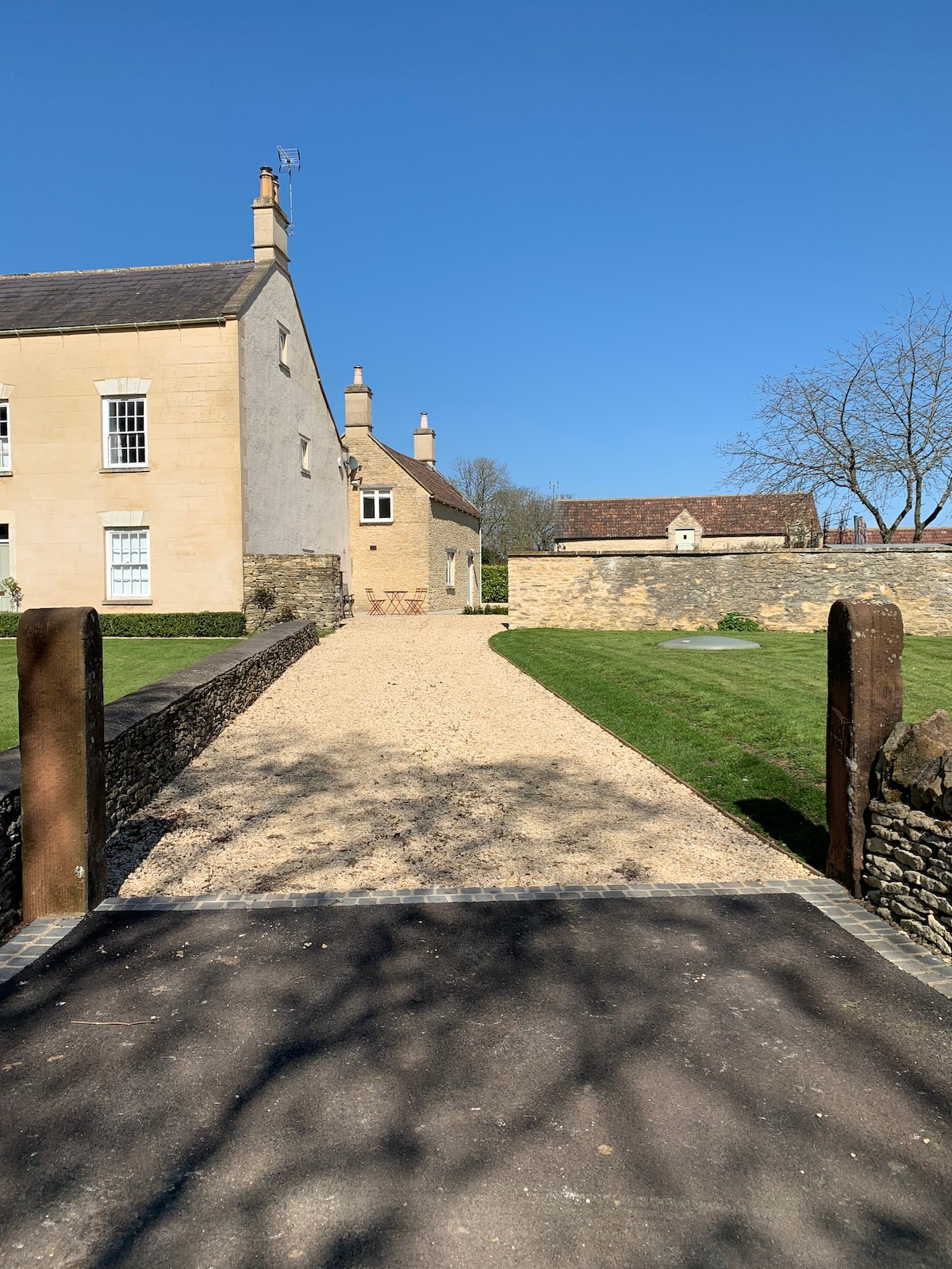 A gravel driveway leads up to the character farmhouse and its annex, framed by stone walls and lush green lawns. The clear blue sky provides a bright backdrop, while the charming building reflects rustic architectural features characteristic of the area.