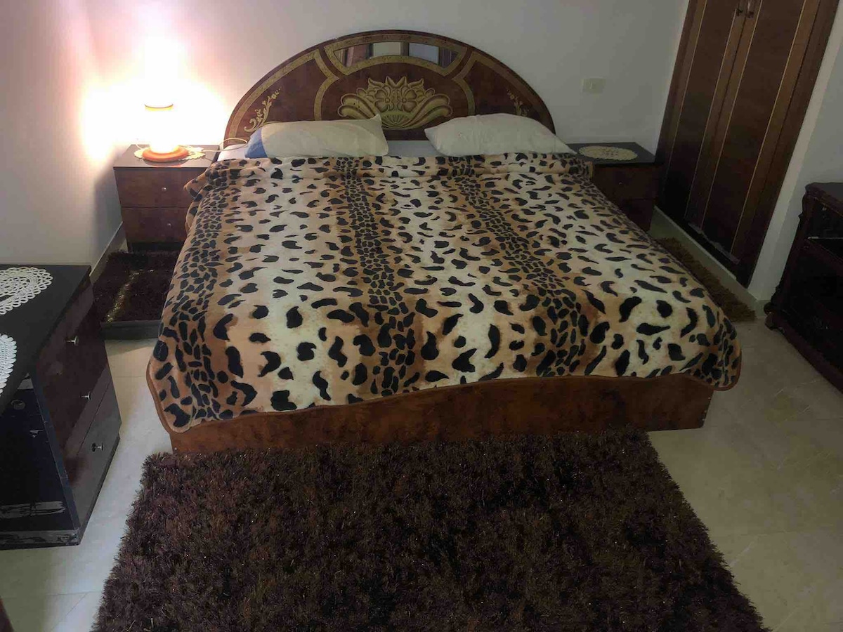 A spacious bed is covered with a patterned blanket featuring a brown and black leopard print. Fruitwood nightstands with lamps are positioned on either side of the bed. The floor is carpeted in a soft brown rug, contributing to a cozy ambiance in the room.
