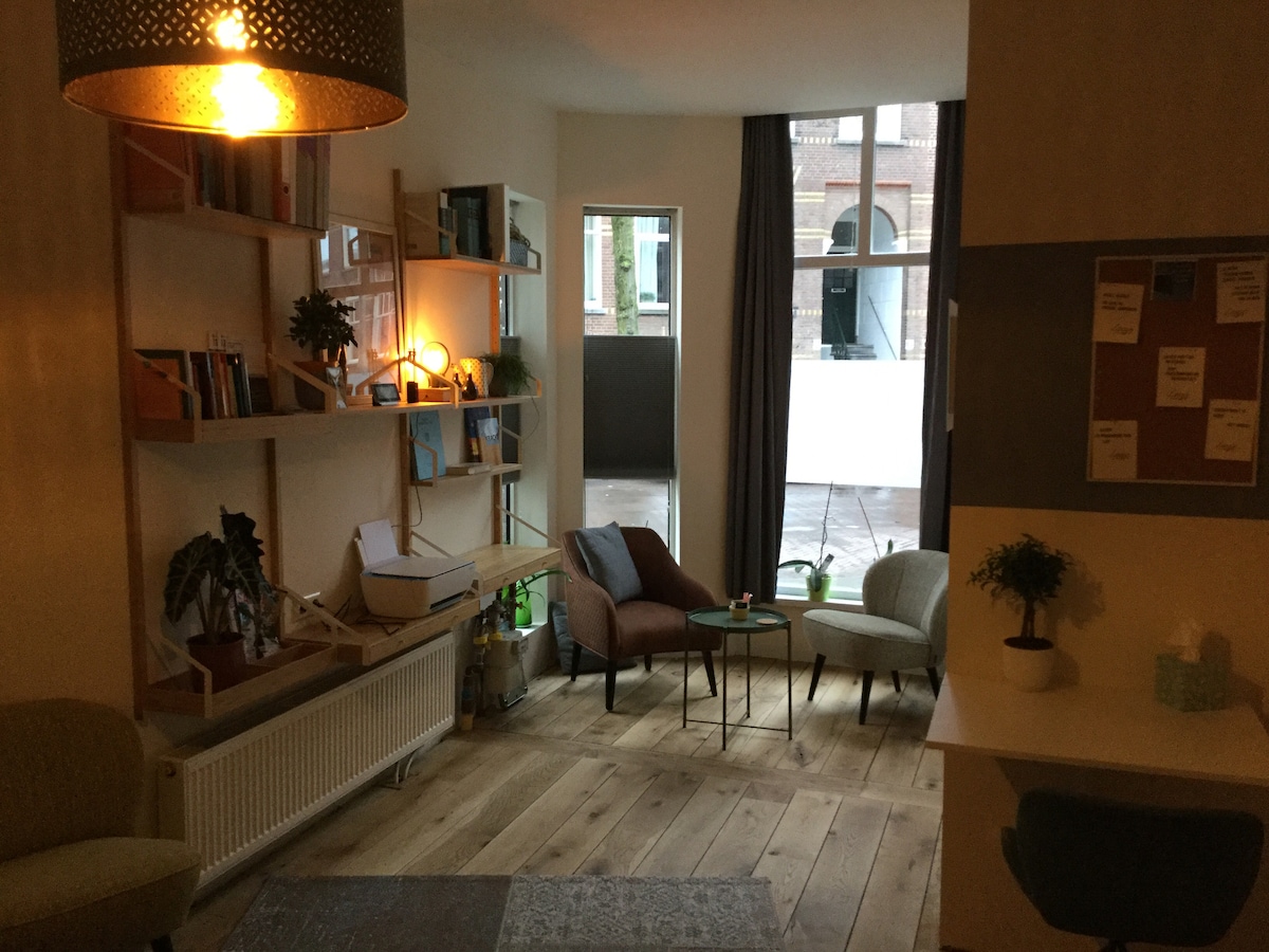 The living area features a combination of light wood flooring and a cozy seating arrangement with two armchairs beside a small table. Shelves line the wall, filled with books and decorative plants. Large windows allow natural light to fill the space, creating a welcoming ambiance.
