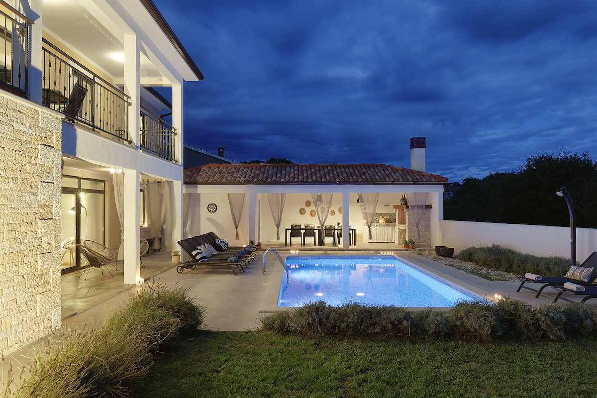 A serene outdoor space is highlighted by a well-lit pool, surrounded by loungers and a dining area. The two-story villa stands in the background, with a covered terrace providing additional seating. Lush greenery and gentle lighting enhance the inviting atmosphere.
