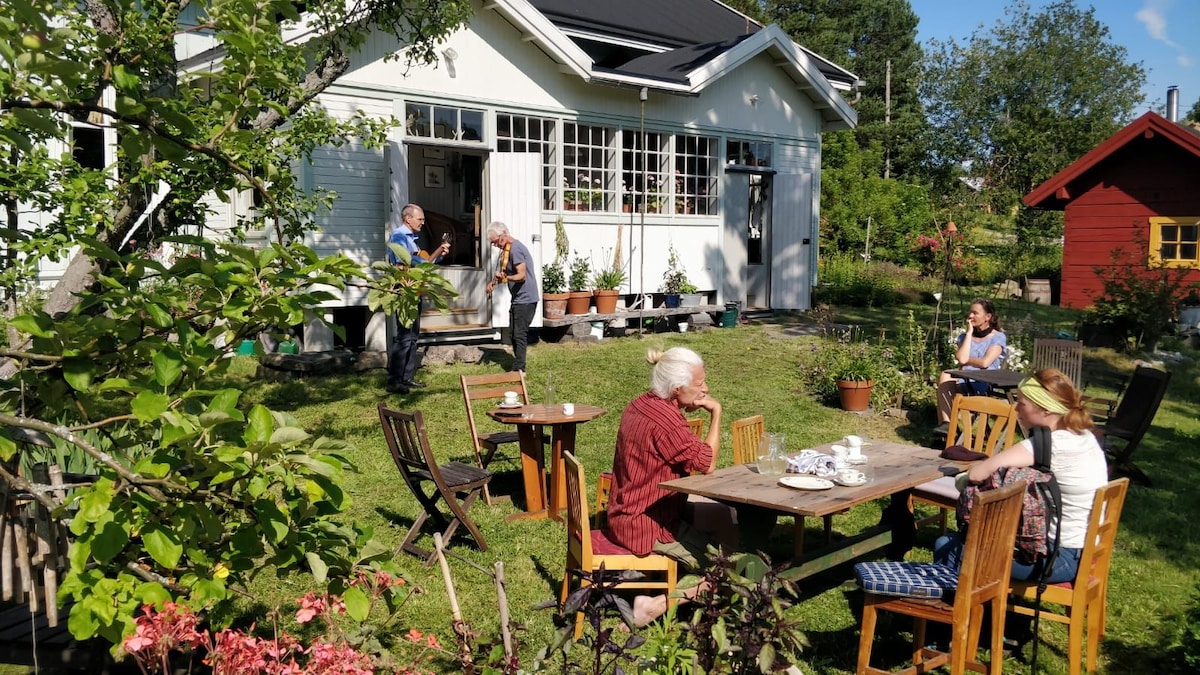 A lush garden setting features several wooden tables and chairs arranged on the grass, inviting social gatherings. A white house with large windows is seen in the background, complemented by green plants and trees. Some guests are engaged in conversation amidst the serene outdoor atmosphere.