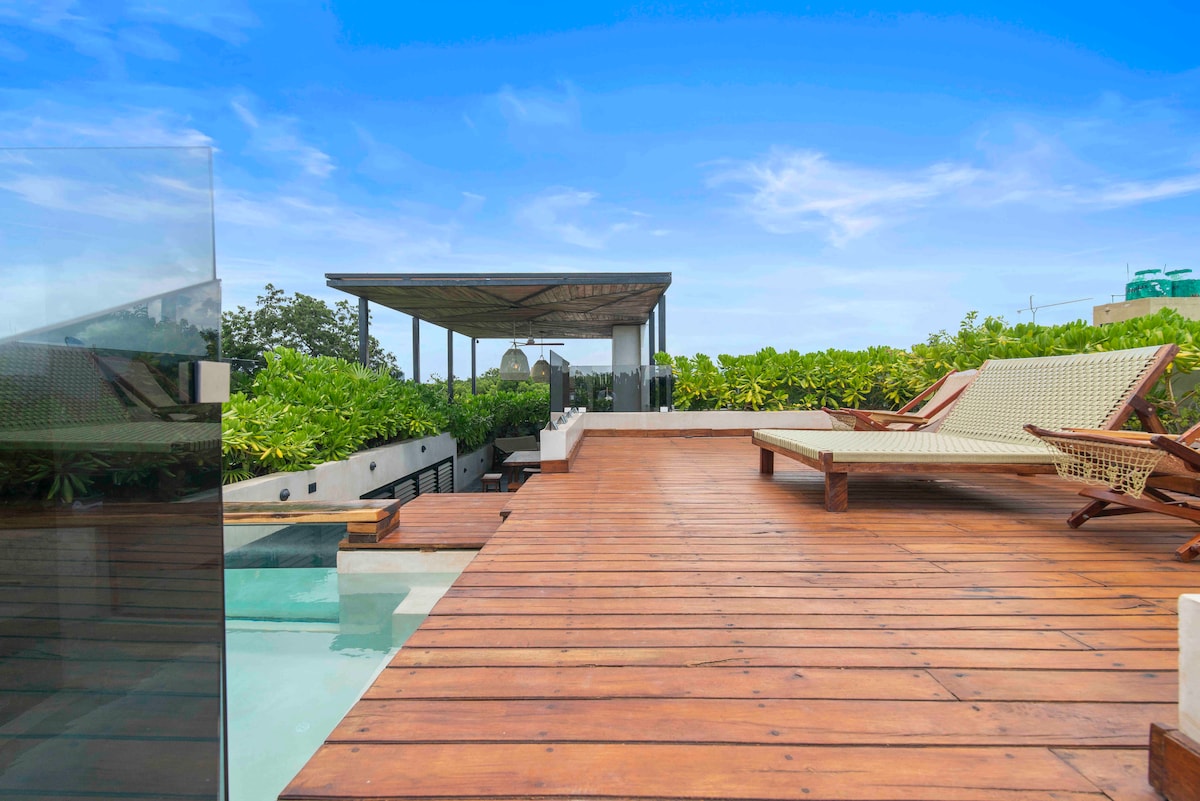 A wooden deck is highlighted with lounge chairs arranged for relaxation. Lush greenery surrounds the area, providing a private atmosphere. A small rooftop pool can be seen to one side, adding to the serene environment.