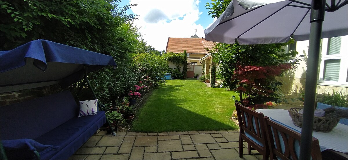 A neatly landscaped garden is visible, featuring a manicured lawn surrounded by various plants and flowers. Comfortable seating is provided under a large umbrella, with a wooden dining set positioned nearby. The serene setting is complemented by greenery and a clear sky.