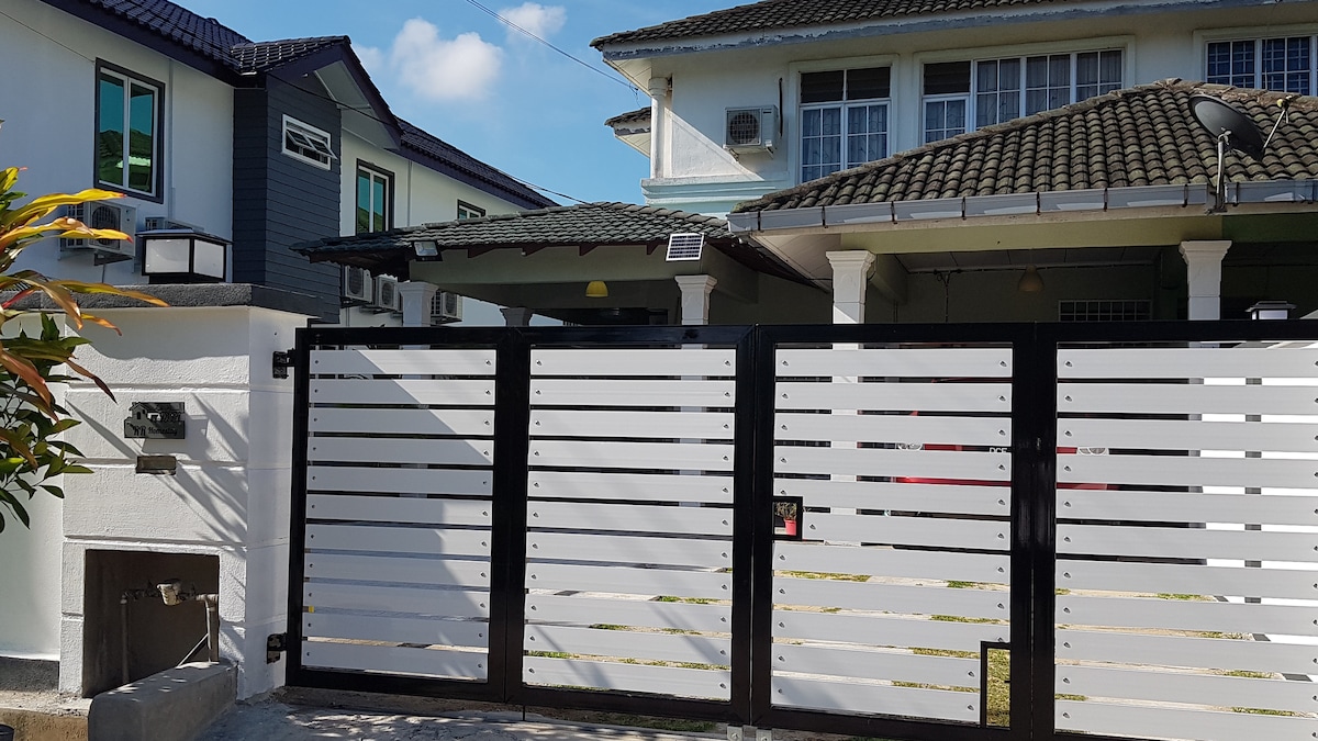 A modern entrance gate features horizontal slats painted in white, framed by a sleek black border. Visible behind the gate is a semi-detached bungalow with a tiled roof and multiple windows, set against a clear blue sky.