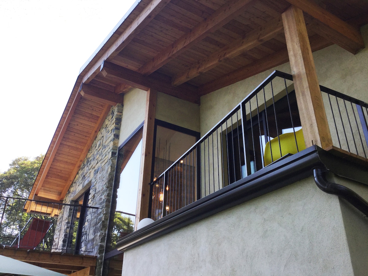 The exterior features a wooden balcony supported by sturdy beams, offering a glimpse of the modern design elements. Large windows provide ample natural light, and a combination of stone and stucco walls adds texture to the facade.
