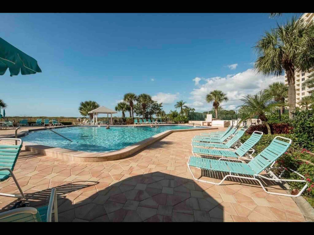 A communal pool area is shown with turquoise lounge chairs arranged around the water’s edge. Palm trees and landscaped greenery provide a tropical setting. Shade is offered by umbrellas, and a gazebo is visible in the background against a clear blue sky.
