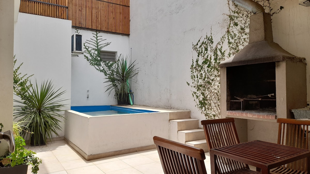 An outdoor space is highlighted by a small pool and a barbecue area. Surrounding foliage provides a touch of greenery, while a patio seating set is positioned for relaxation. Light-colored tiles create an open, airy feel in this inviting area.