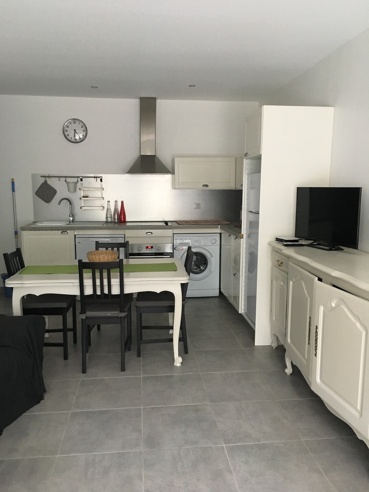 A modern kitchen and dining area are visible, featuring a compact table with four chairs. Appliances include a washing machine and a refrigerator, along with a stove and a kitchen sink against the wall. A clock hangs above, and a television is placed on a sideboard.