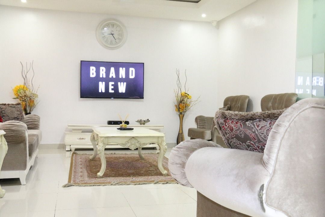 A bright living room is featured, with modern furniture including plush armchairs and a central coffee table. A large flat-screen television is mounted on the wall, with decorative vases and a soft area rug enhancing the overall elegance.
