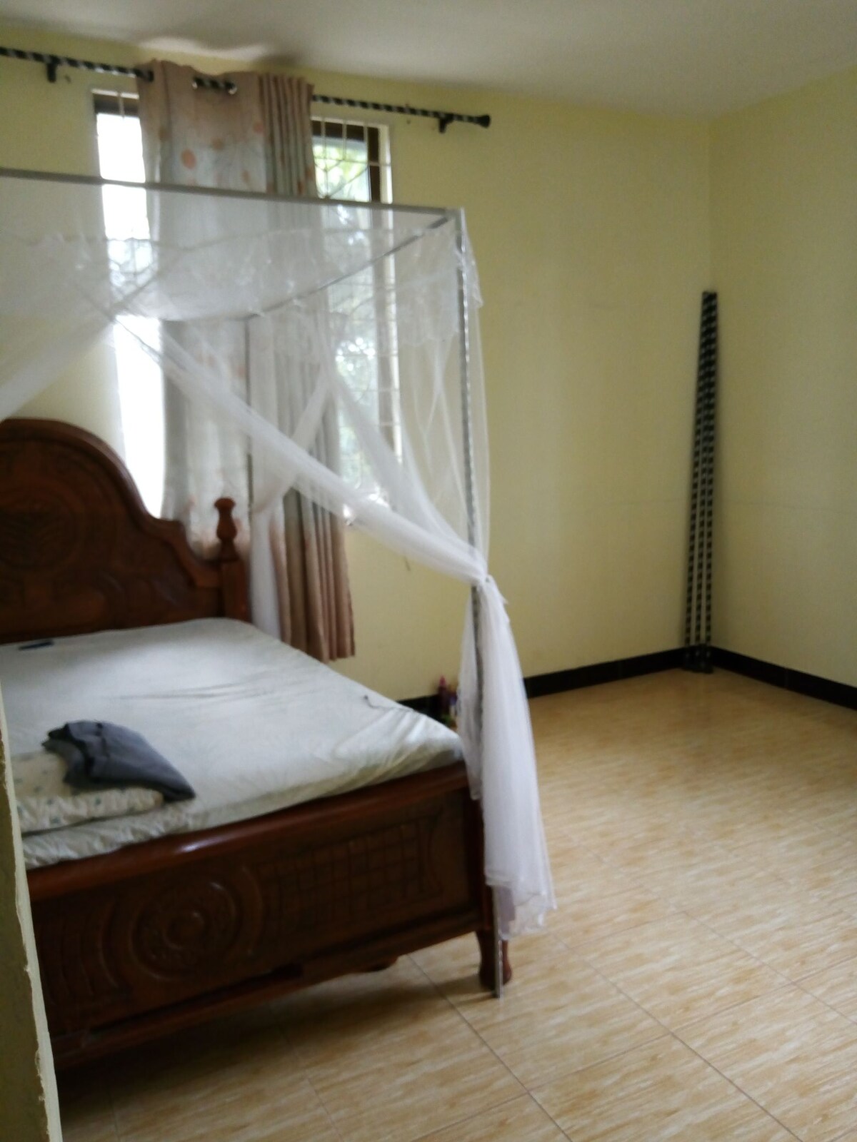A spacious bedroom features a wooden bed with a decorative canopy. Natural light enters through large windows covered by sheer curtains. The floor is tiled, and minimal furnishings are present, creating an open and airy ambiance.