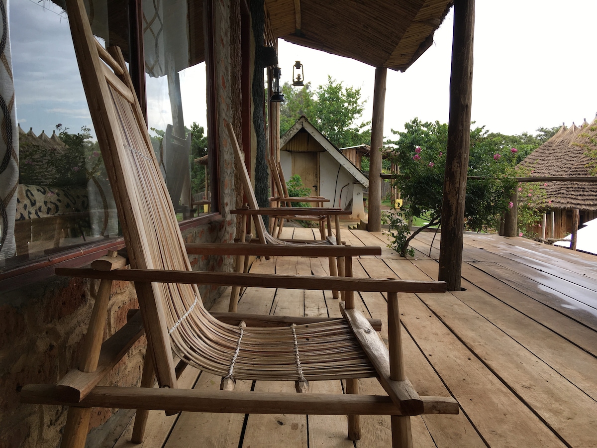 Two wooden lounge chairs are positioned on a covered porch, facing the scenic landscape. The natural wooden surface of the porch complements the rustic style of the nearby structures, framed by lush greenery and hints of vibrant flowers.