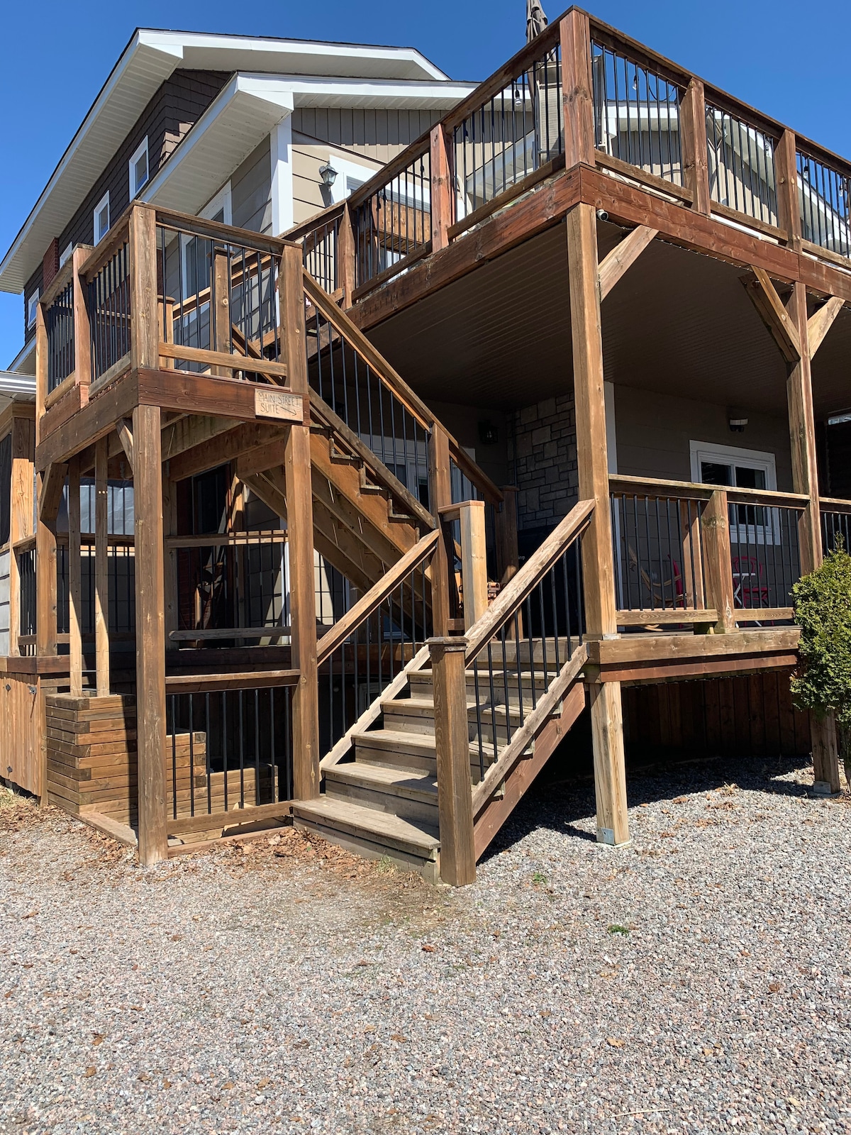 A multi-level wooden deck is depicted, featuring sturdy railings and steps leading to an upper entrance. The deck's rustic design complements the exterior of the house, with gravel covering the ground beneath. Natural light illuminates the outdoor space, enhancing its inviting character.