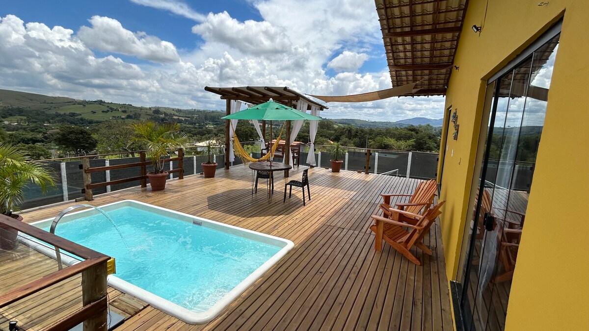An outdoor space features a wooden deck with a heated pool and built-in whirlpool. A pergola provides shade over a dining area, which includes a table and chairs. Lush greenery and scenic mountain views are visible in the background, enhancing the tranquil setting.