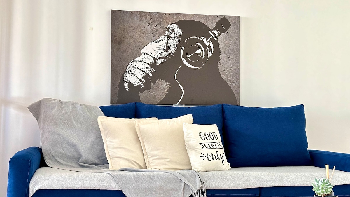 A blue sofa is displayed in a living area, complemented by a soft gray throw and decorative pillows. A striking artwork of a monkey with headphones hangs on the wall, adding an artistic touch to the space.