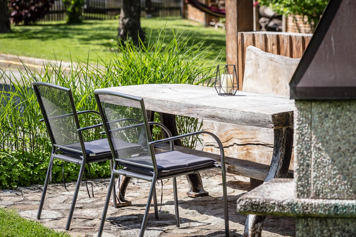 A rustic wooden table is paired with two metal chairs, set on a stone patio surrounded by lush greenery. A geometric lantern is placed on the table's surface, enhancing the outdoor dining space's inviting atmosphere.