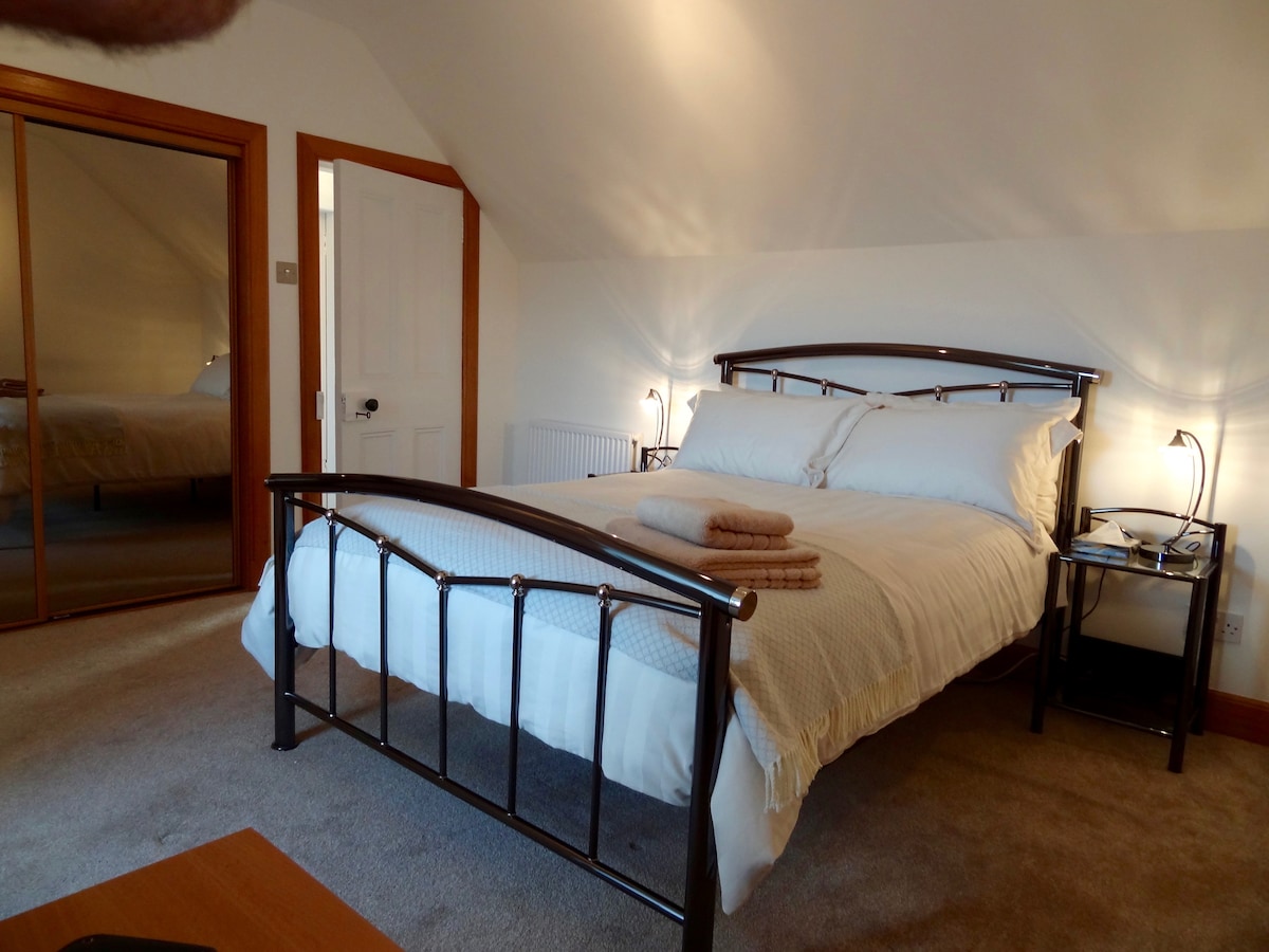 A well-lit bedroom features a black metal frame bed with neatly arranged bedding and towels. A full-length mirror reflects the cozy space, while bedside lamps provide additional lighting. The room is carpeted, enhancing comfort, with a wooden nightstand beside the bed.