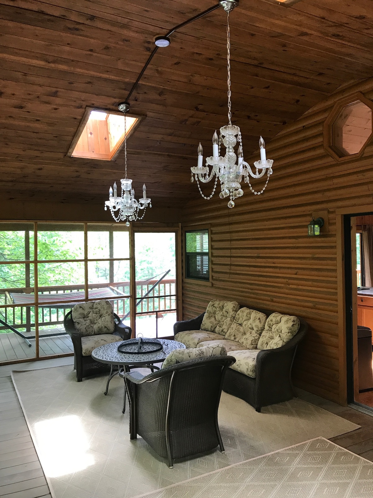 An enclosed seasonal room features comfortable seating arranged around a round table. Crystal chandeliers hang from a wooden ceiling, and natural light streams through a skylight and ample windows, enhancing the connection with the surrounding greenery.