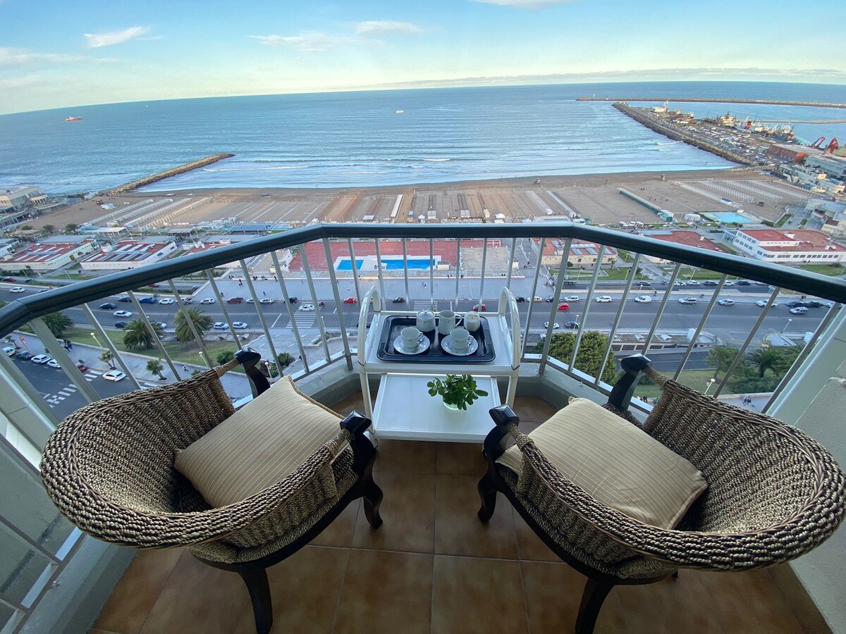 A balcony offers a view of the sea and beach below, featuring two woven chairs with cushions. A small white table holds dinnerware and a potted plant, while the horizon stretches in the background, enhancing the serene coastal setting.