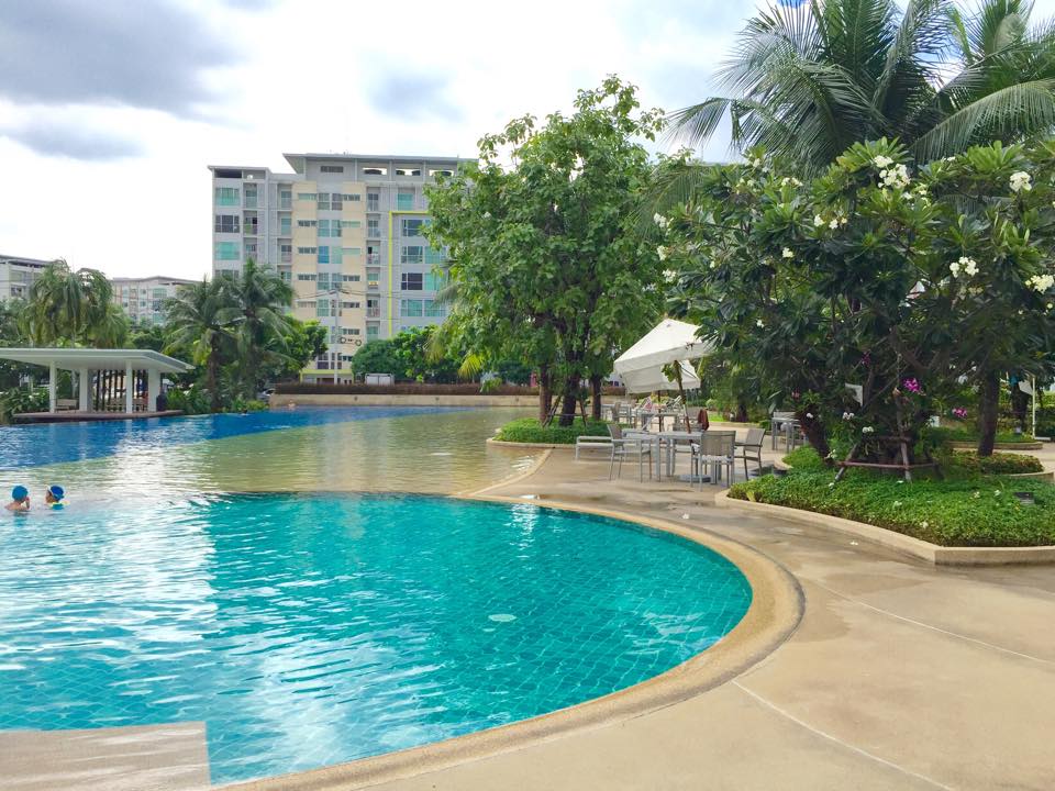 The image depicts a large swimming pool surrounded by lush greenery, including palm trees and flowering plants. Sun loungers and shaded seating areas are visible, inviting relaxation. The pool area is separated into shallower and deeper sections, with residents enjoying the water on a calm day.